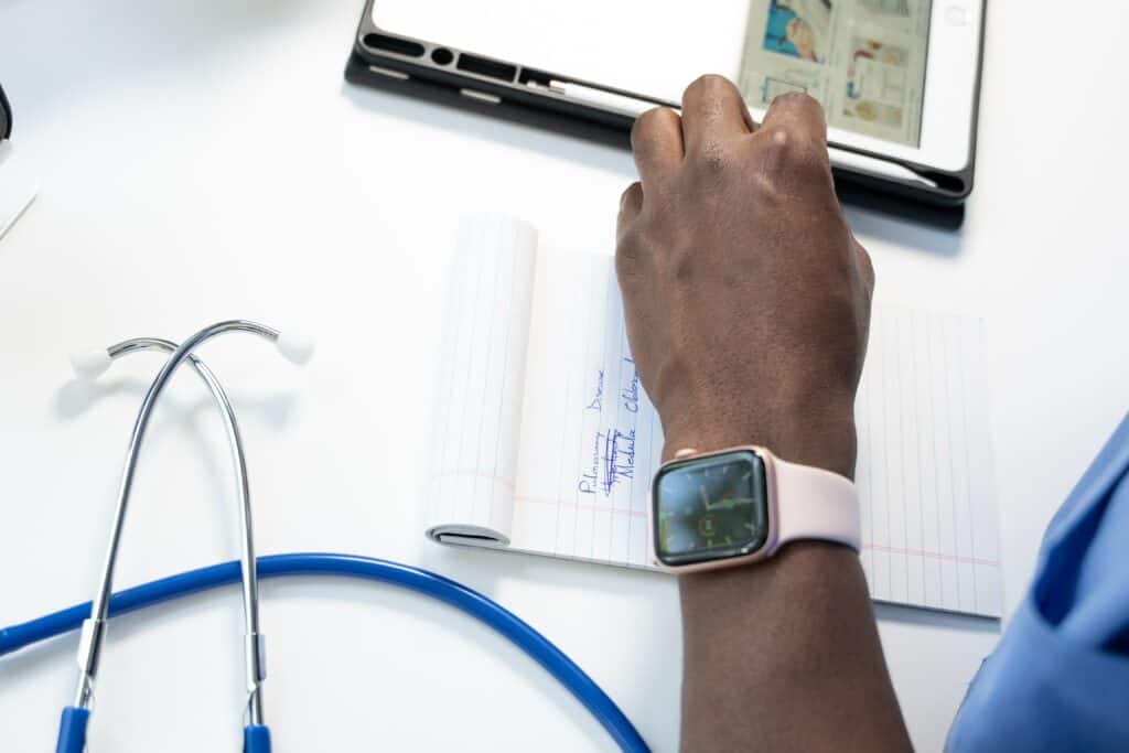 Left arm of a healthcare professional with a smart watch on resting on a table with a computer, stethoscope and notepad