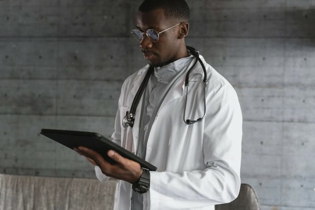 A Black doctor in a gray room checks a tablet for patient messages as part of automated medical answering services. He wears glasses, a white lab coat and a stethoscope around his neck.